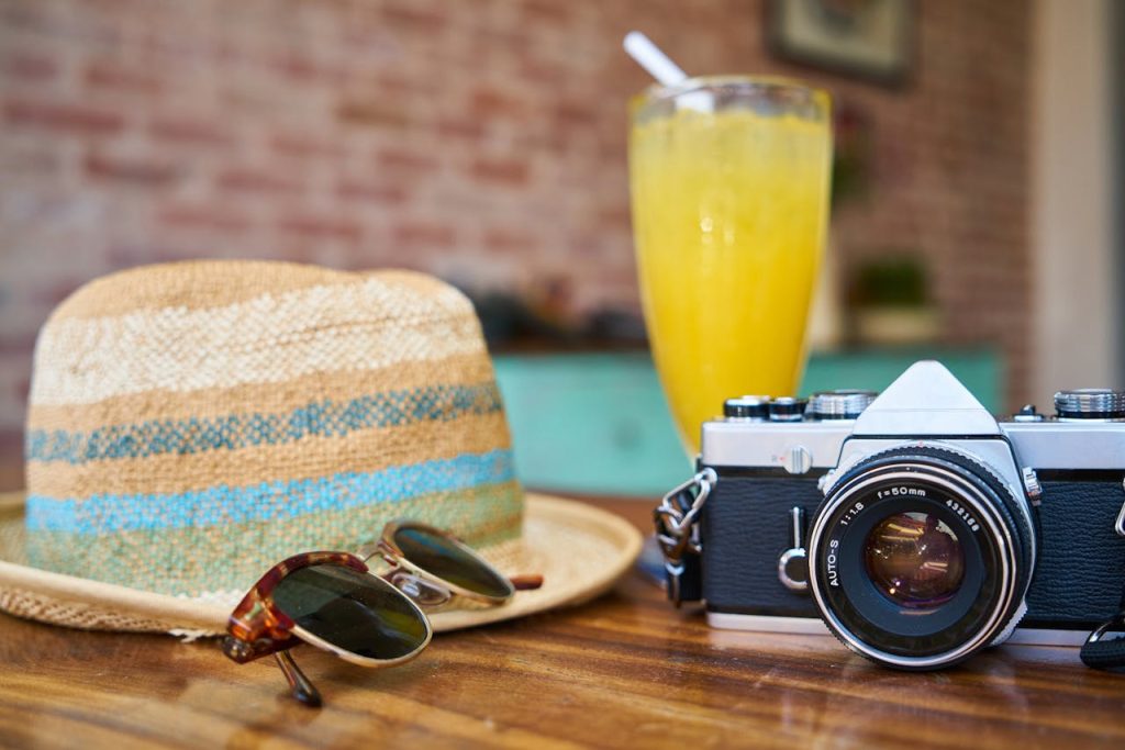 lunettes de soleil, chapeau de paille, appareil photos sont posés sur une table avec un cocktail jaune. Ambiance vacances